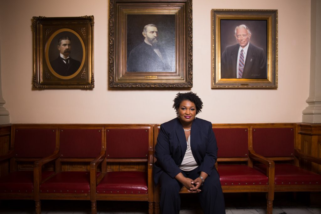 Image of Stacey Abrams sitting in a red leather chair in front of pictures of past Georgia governors at the state house in Atlanta, Georgia. 
